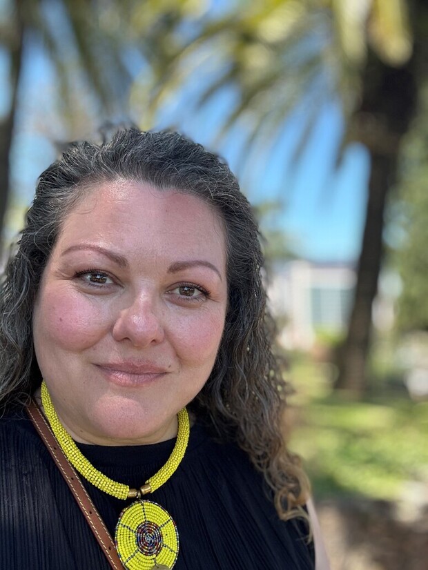 A woman with curly hair smiles gently, wearing a yellow beaded necklace with a circular pendant, standing outdoors with palm trees and a bright blue sky behind her.